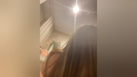A blurry video of a woman with long brown hair, seen from behind, standing in a dimly lit bathroom with a ceiling light creating a lens flare effect.