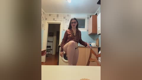 Video of a young woman with glasses, long brown hair, and a maroon shirt, sitting on a wooden chair in a modest, tiled kitchen with light blue walls.