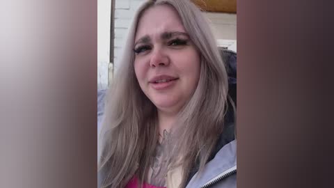 Video of a middle-aged woman with long, straight, platinum blonde hair, light skin, and a faint smile. She wears a gray jacket over a pink top. Background is blurred, with hints of a house exterior.