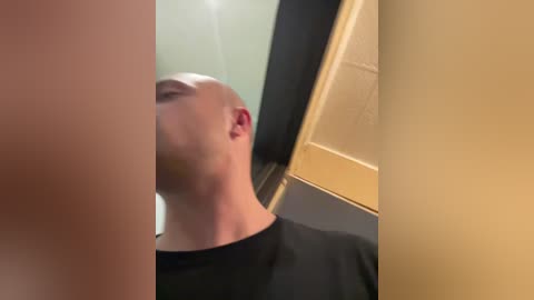 Video of a bald, fair-skinned man with a short beard, wearing a black shirt, standing in a modern elevator with beige walls and a glass door.