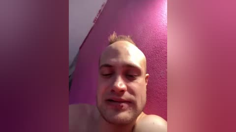 Video of a bald, light-skinned man with a small tuft of hair on his head, standing in front of a pink wall. His eyes are closed, and he has a neutral expression.