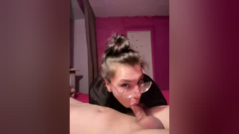 A video depicts a woman with short, dark hair in a bun, wearing glasses, performing oral sex on a man in a room with pink walls and curtains.