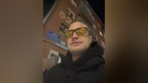 Video of a bald, light-skinned man with a mustache wearing yellow sunglasses, black hoodie, and a white iPhone in his hand, taken at night against a blurred, dimly lit background.