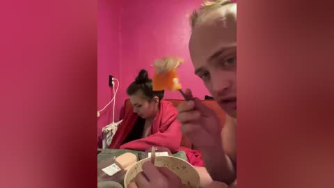 Video of two young girls with blonde pigtails and red dresses, sitting on a bed with pink walls, playing with a toy.