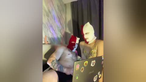 Video of a muscular, tattooed man in a red mask and white hood, sitting shirtless on a bed with a laptop, surrounded by colorful, abstract wall art.