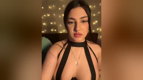 Video of a light-skinned woman with dark hair, wearing a black halter top, adorned with a choker necklace. Background features a warm, dimly lit room with fairy lights.