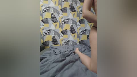A video of a person, partially visible, lying on a bed with gray bedding, under a mustard-yellow, gray, and black leaf-patterned wallpaper. The image is soft-focused.