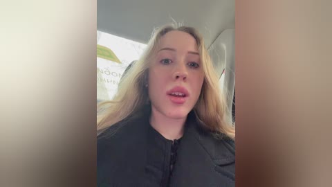 Video of a Caucasian woman with long blonde hair, wearing a black jacket, standing inside a car with a blurred background.
