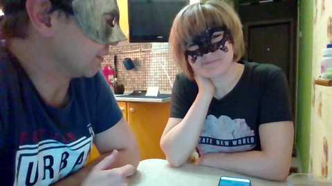 Video of a young man in a baseball cap and a woman in a black mask, both wearing t-shirts, sitting at a kitchen table with a tiled backsplash and cabinets.