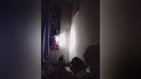 A dimly lit video of a child lying on a bed with a colorful blanket, in a small, cramped room with a single light bulb illuminating the corner.