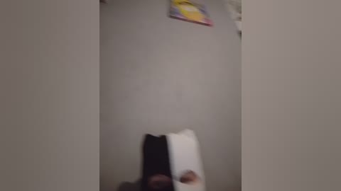 A blurred video of a narrow, beige-walled bathroom with a white towel and a partially visible person, possibly in a seated position, at the bottom. A yellow, red, and white sign hangs above, partially out of frame.