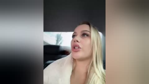 Video of a blonde woman with fair skin, wearing a white robe, in a dimly lit room. Her expression is contemplative, with slightly parted lips. The background shows blurred, indistinct objects.