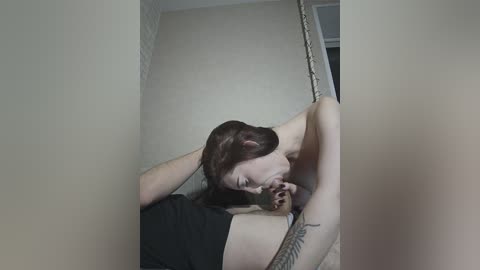 Video of a nude woman with long dark hair, wearing a black t-shirt, leaning against a beige wall with a visible tattoo on her arm.