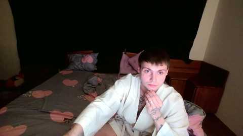 A video of a young man with short brown hair in a white bathrobe, sitting on a bed with a pink heart-patterned blanket, in a dimly lit room.