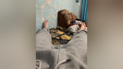 A video of a child lying on a patterned quilt, wearing a gray hoodie and pajama bottoms, against a blue wallpaper with floral designs. The child's hands are near their head.