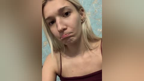 A video of a young woman with fair skin and straight blonde hair, wearing a maroon spaghetti strap top, pouting with a slightly crooked nose, against a blue floral-patterned background.