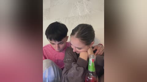 A video of two young people, a boy and a girl, sitting close, with the girl leaning on the boy's shoulder, wearing a pink shirt and brown jacket, while the boy wears a gray shirt. A soda bottle is in the foreground.
