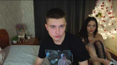 Video of a young man with short brown hair, wearing a Star Wars T-shirt, sitting on a bed in a dimly lit room with a woman in lingerie.