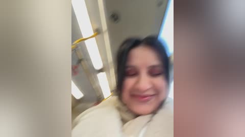 Video of a smiling woman with dark hair, wearing a white coat, standing in an elevator with fluorescent lights and a yellow handrail, blurred background.