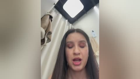 A candid video of a young woman with long brown hair, wearing makeup, looking up in a bathroom with beige walls, a white shower curtain, and a small window.