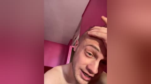 A close-up video of a young, fair-skinned, clean-shaven man with short brown hair, wearing a white shirt, and sitting in a room with pink walls. He looks tired and rubs his forehead with his right hand.