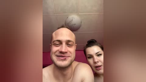 Video of a bald, shirtless man with a smug expression and a woman with a bun, both in a pink bathroom with a ceiling light fixture.