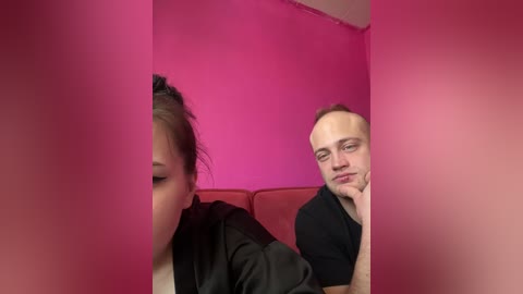 Video of a young Asian woman with dark hair in a bun, leaning against a red wall, and a bald, middle-aged man with fair skin, wearing a black shirt, both appearing bored.