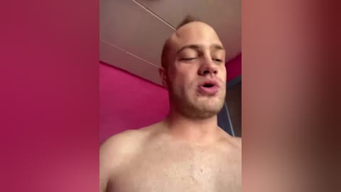 Video of a bald, light-skinned, shirtless man with closed eyes, mouth slightly open, standing against a pink wall with a ceiling light.