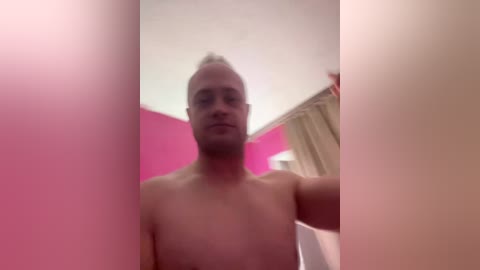 Video of a shirtless, balding, muscular man with a light skin tone, standing in a pink-walled room. His right hand is raised, possibly holding a curtain. The background includes a beige curtain and a ceiling light.