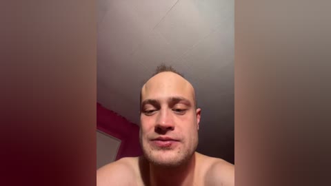 Video of a bald, light-skinned man with a trimmed beard, wearing no shirt, standing in a room with a pink wall and white ceiling.