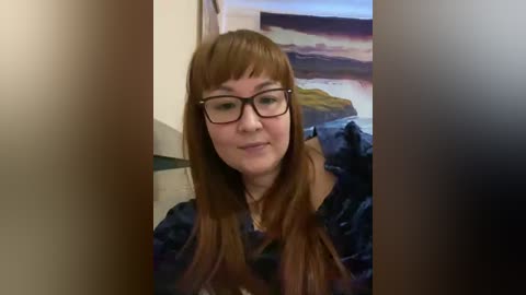 Video of a Caucasian woman with long auburn hair, wearing glasses and a dark-patterned blouse, standing indoors. Background features a blurred, abstract painting of a sunset.