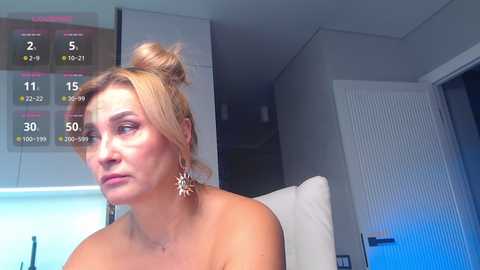 Video of a blonde woman with light skin, wearing a sunflower earring, and a distressed expression. She's indoors with a calendar showing May 2021 and a door in the background.