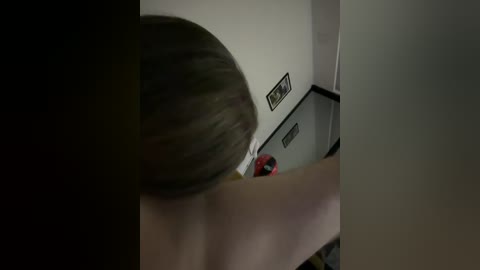 Video of a woman with short, dark hair, possibly in her early 20s, standing in a dimly lit, cramped room. She's wearing a white t-shirt and holding a red object. The walls are white, and there's a framed photo on the right.