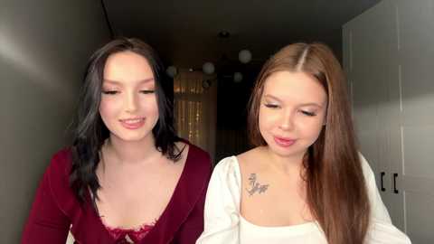 Video of two young women with fair skin, one with black hair and a maroon top, the other with long brown hair and a white top, smiling and standing in a narrow hallway.