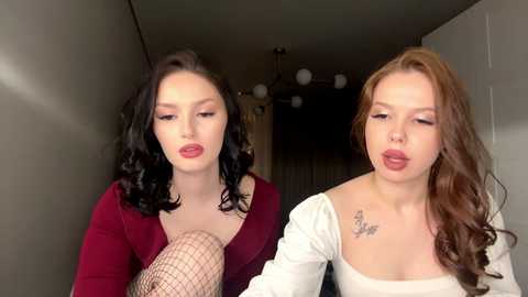 Video of two young women with fair skin, one with dark wavy hair, the other with light brown curls, in a dimly lit hallway. They wear red lipstick, fishnet stockings, and off-shoulder tops, kissing passionately.