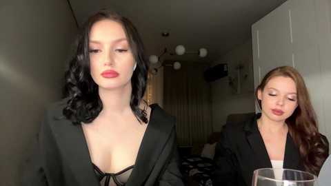 Video of two women in a dimly lit room. One, with long black hair and red lipstick, wears a black blazer over a black bra, standing in the foreground. The other, with long auburn hair, light makeup, and a black blazer, sips from a glass in the background.