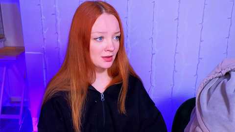 Video of a young, fair-skinned woman with long, straight red hair, wearing a black robe, sitting indoors with string lights and a beige blanket in the background.