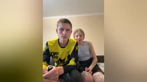Video of a young, pale-skinned man with short, light brown hair, wearing a yellow and black soccer jersey, sitting on a bed in a plain room. Behind him, a fair-skinned woman with blonde hair, wearing a white tank top and black skirt, sits with a neutral expression.