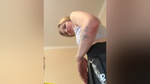 A video shows a blonde woman with a medium build, wearing a white tank top and black shorts, bending over, revealing a tattoo on her upper arm. The background features a beige wall and a ceiling with white trim.