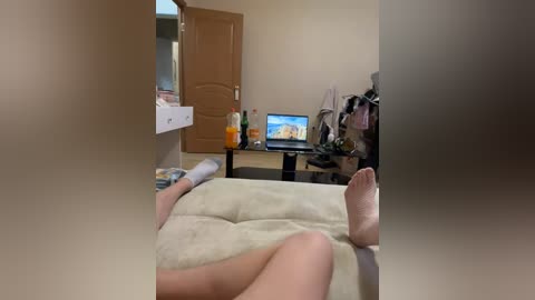 A video showing a beige bed with a woman's feet visible, a closed door, a desk with two computer screens, and a chair, creating a cozy, casual bedroom scene.