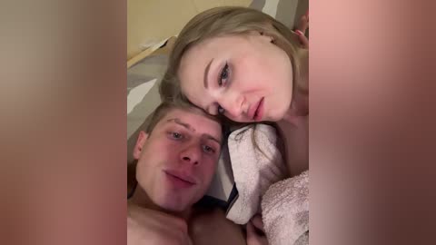 Video of a couple in bed, a blonde woman with fair skin and a shirtless man with light skin, both smiling, slightly blurred.