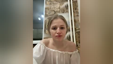 A video of a young Caucasian woman with light brown hair styled in a bun, wearing a white off-the-shoulder blouse, standing in front of a mural depicting ancient architecture.