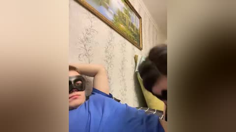 Video of a man in a blue shirt and black mask, arms raised, standing in a room with a white floral wallpaper, framed painting, and yellow cushion.