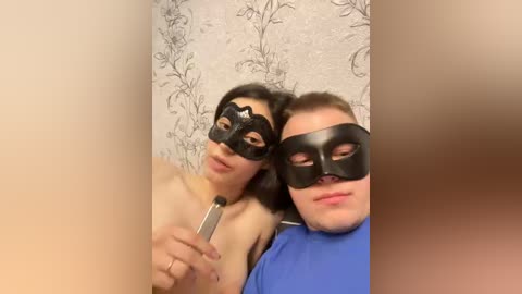 A video of two people wearing black masquerade masks, one with long dark hair, the other with short brown hair. They stand against a wallpaper with a floral pattern.