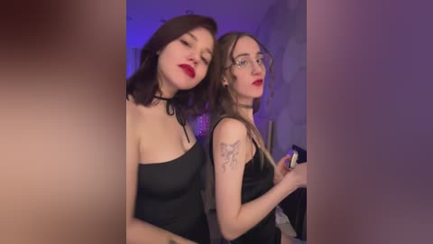 Video of two young women with pale skin, dark hair, and red lipstick. One wears a black strapless dress, the other a black top. Background features dim lighting and a blurred wall.