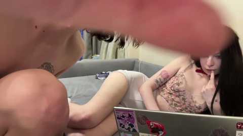 Video of a nude man with a shaved head, partially visible, standing in front of a mirror. A tattooed woman with long dark hair, wearing floral lingerie, sits on a gray couch in a cluttered room.