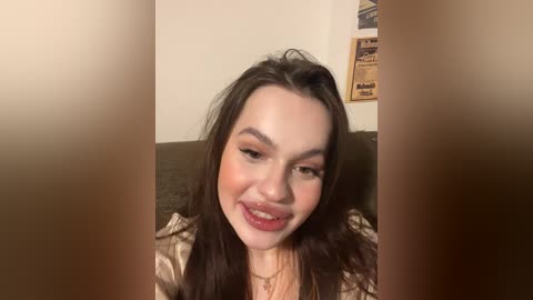 Video of a young woman with long, dark hair, fair skin, and light makeup, smiling widely, set against a beige wall with framed pictures.