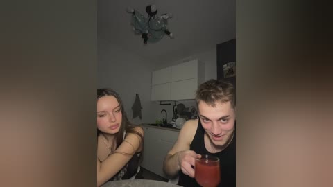 Video of a young Caucasian couple in a dimly-lit kitchen; man with short hair, tank top, holding a glass of red drink; woman with long hair, tank top, looking contemplative.