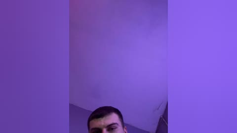 A video of a man with short dark hair, partially visible, standing in a dimly lit room with a purple hue. The background is a plain, off-white wall with a faint shadow on the right.