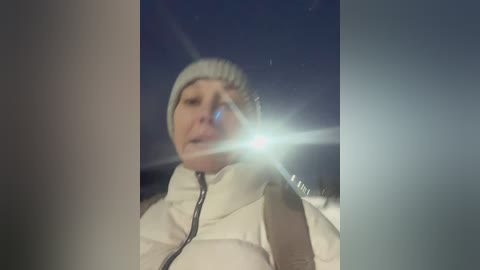 Video of a middle-aged woman with fair skin and light hair, wearing a beige winter coat and a knitted beanie, driving at night with a bright light reflecting off the car's windshield.